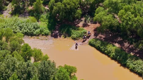 Cowboy-adventure-group-crosses-muddy-river-on-horseback,-wading-through-unknown-territory,-aerial-orbit