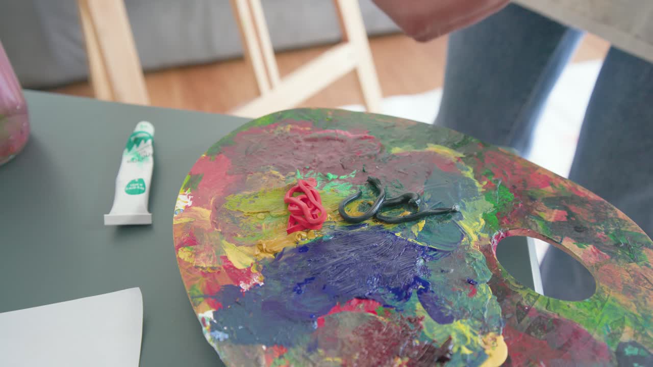 Mani che spremono un tubetto di vernice rossa, lo tappano e mescolano il rosso con i verdi su una tavolozza per dipingere.