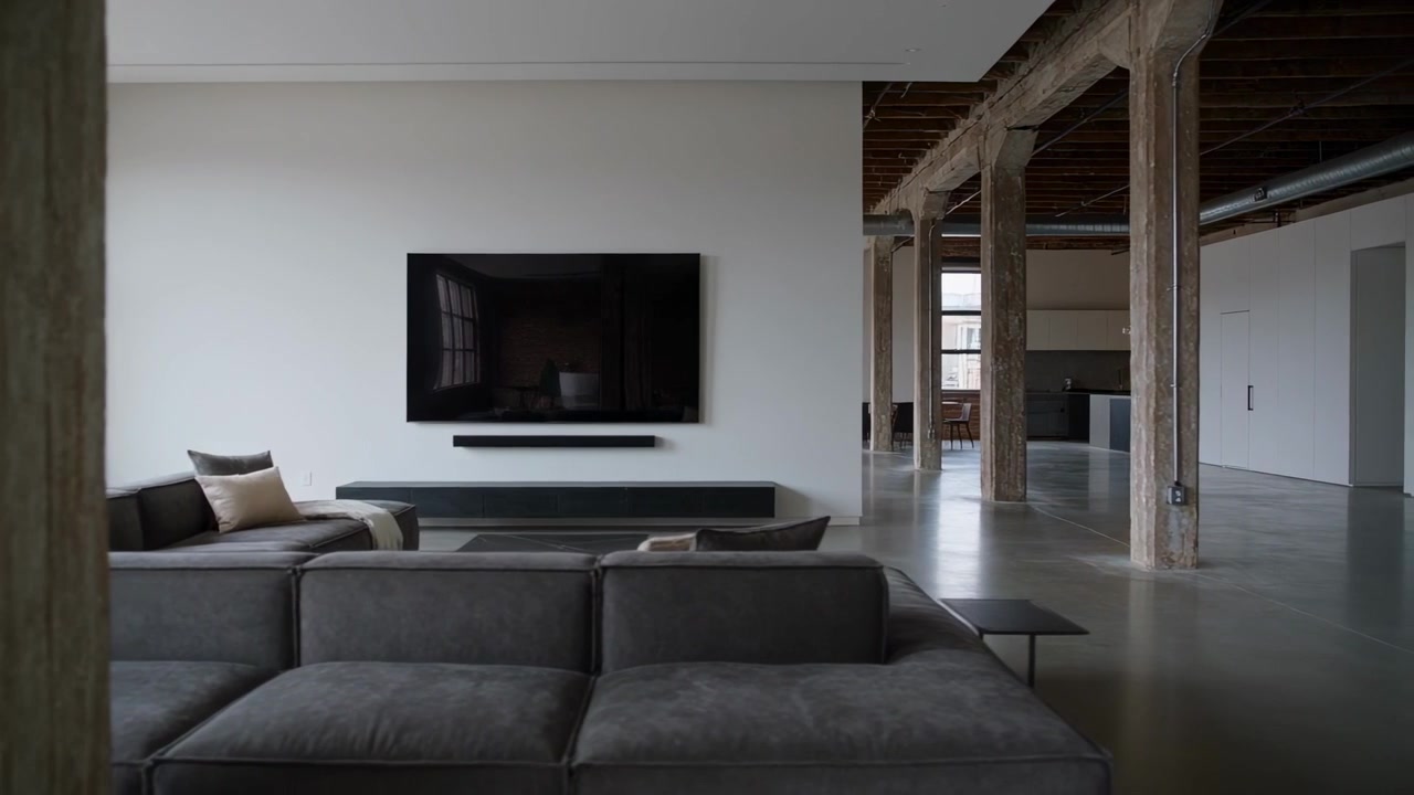 Cámara panorámica comenzando desde la derecha, revelando un loft con sofá seccional gris, televisor y columnas expuestas.