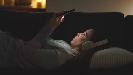 Woman Wearing Wireless Headphones Lying On Sofa At Home At Night Streaming Or Looking At Online Content On Mobile Phone 5