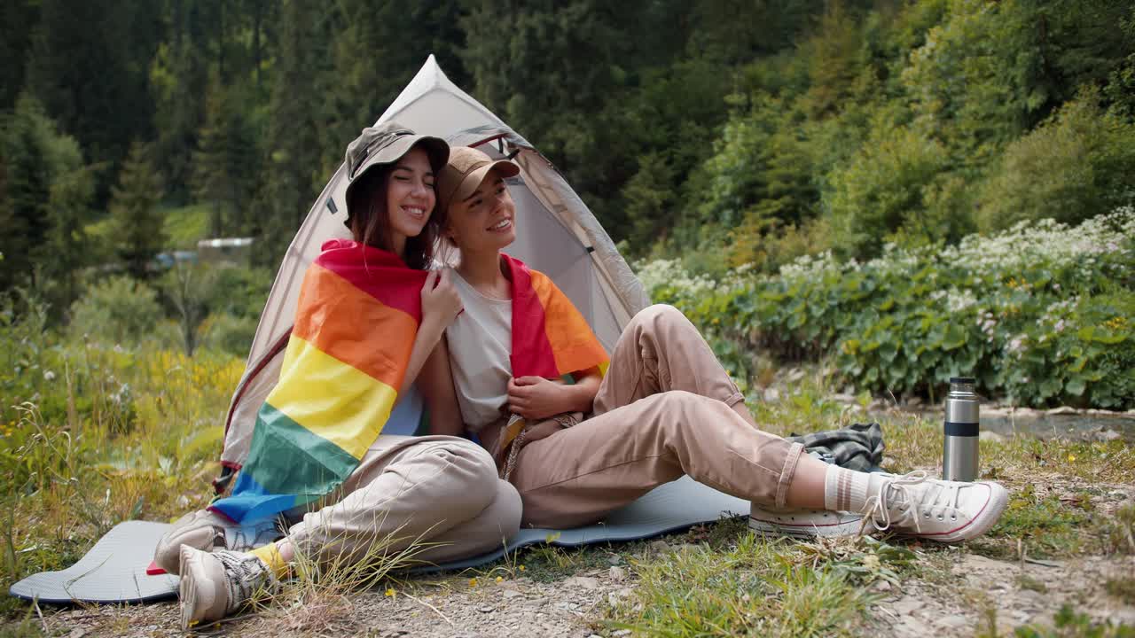 una ragazza bionda e una ragazza lesbica bruna sono sedute l'una accanto all'altra avvolte in una bandiera lgbt vicino a una tenda sullo sfondo di una foresta verde