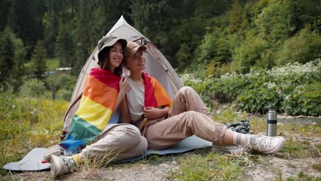 Una-Chica-Rubia-Y-Una-Lesbiana-Morena-Están-Sentadas-Una-Al-Lado-De-La-Otra-Envueltas-En-Una-Bandera-Lgbt-Cerca-De-Una-Tienda-De-Campaña-Con-El-Telón-De-Fondo-De-Un-Bosque-Verde