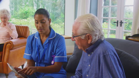 senior couple at home with woman talking to female nurse or care worker using digital tablet