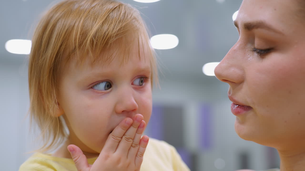 primer plano de madre e hija en una interacción sincera, el niño cubre la boca mientras se relaciona con la madre, quien responde con calidez, iluminación suave y luces borrosas del techo
