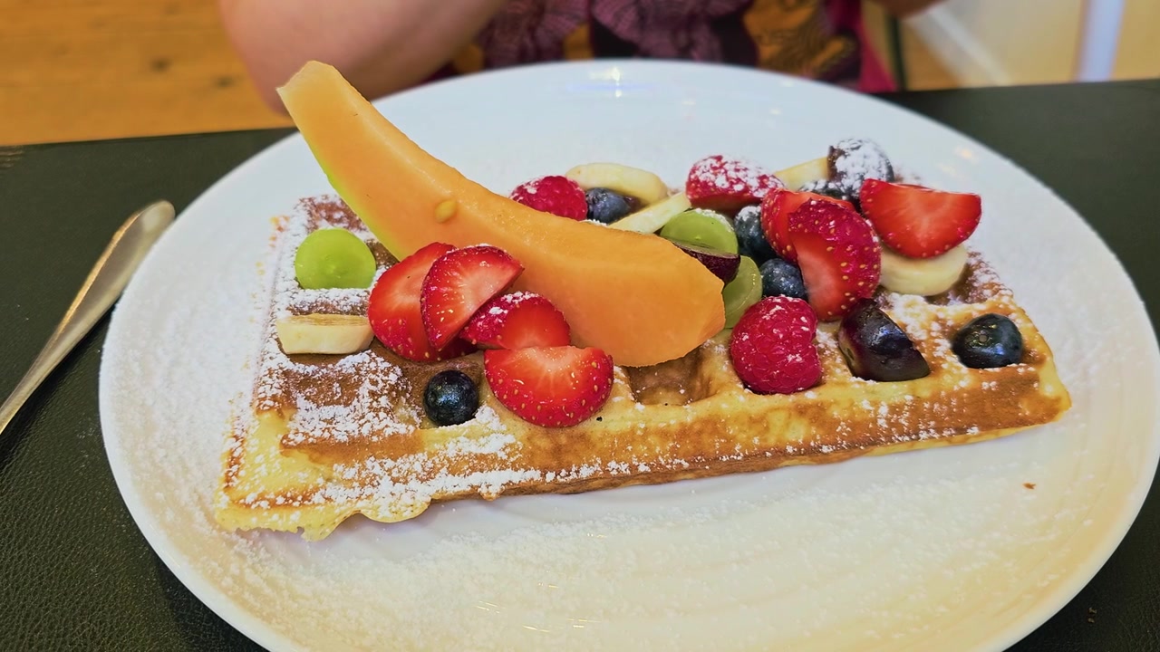 Dessert waffle in stile europeo con frutti di bosco freschi, banana a fette, uva, melone, zucchero a velo e una consistenza croccante e dorata, servito su un piatto bianco su un accogliente tavolo da colazione in un caffè al chiuso.