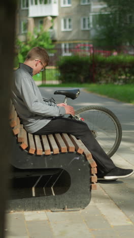 un niño con una camiseta gris y pantalones negros se aleja de su bicicleta estacionada, se sienta en un banco de madera en el parque, se inclina hacia atrás y cruza la pierna, con una vista borrosa de un edificio residencial
