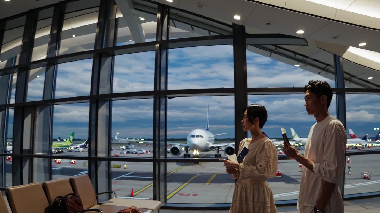 Wide-angle shot of two people at an airport terminal, observing planes through large windows