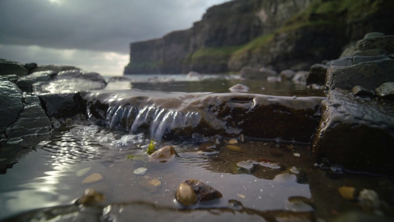 Surging water approaching left pushing over flat wet rock ledge with tidal pool, forming cascade