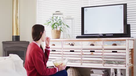 caucasian woman sitting on couch, eating snacks and watching tv with copy space in living room