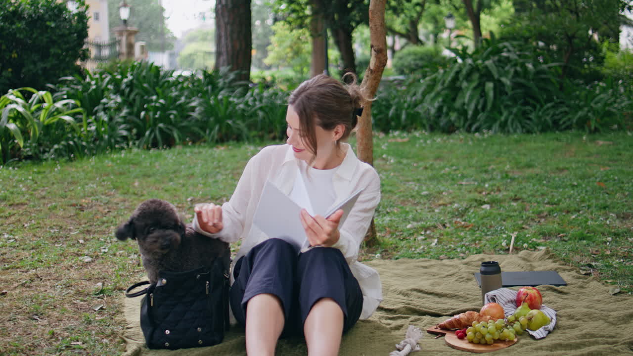 Relaxed girl sitting blanket with dog at park picnic. Happy woman enjoying pet