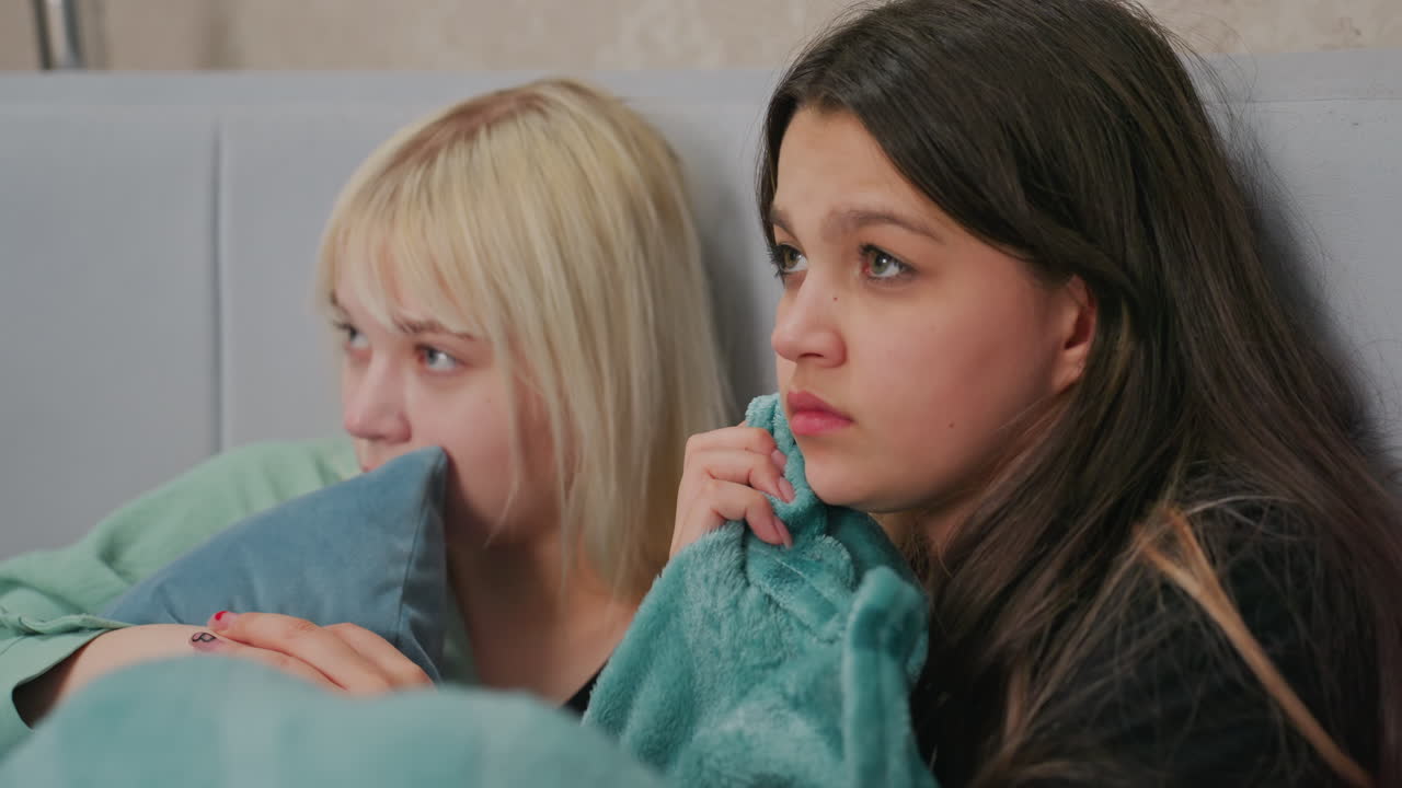young girls sitting on bed watching horror movie with scared expressions, one holding friend hand tightly while biting blanket, both wrapped in blue covers and leaning into each other
