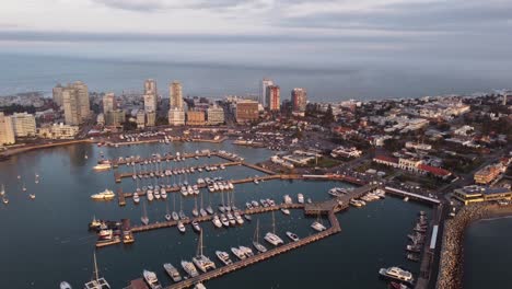 Vuelo-Aéreo-Hacia-Atrás-Que-Muestra-El-Hermoso-Horizonte-De-La-Ciudad-De-Punta-Del-Este-Con-Puerto-Y-Estacionamiento-De-Veleros-Durante-La-Puesta-De-Sol-Dorada---Océano-Atlántico-Sur-En-Segundo-Plano-Cubierto-De-Nubes-Oscuras