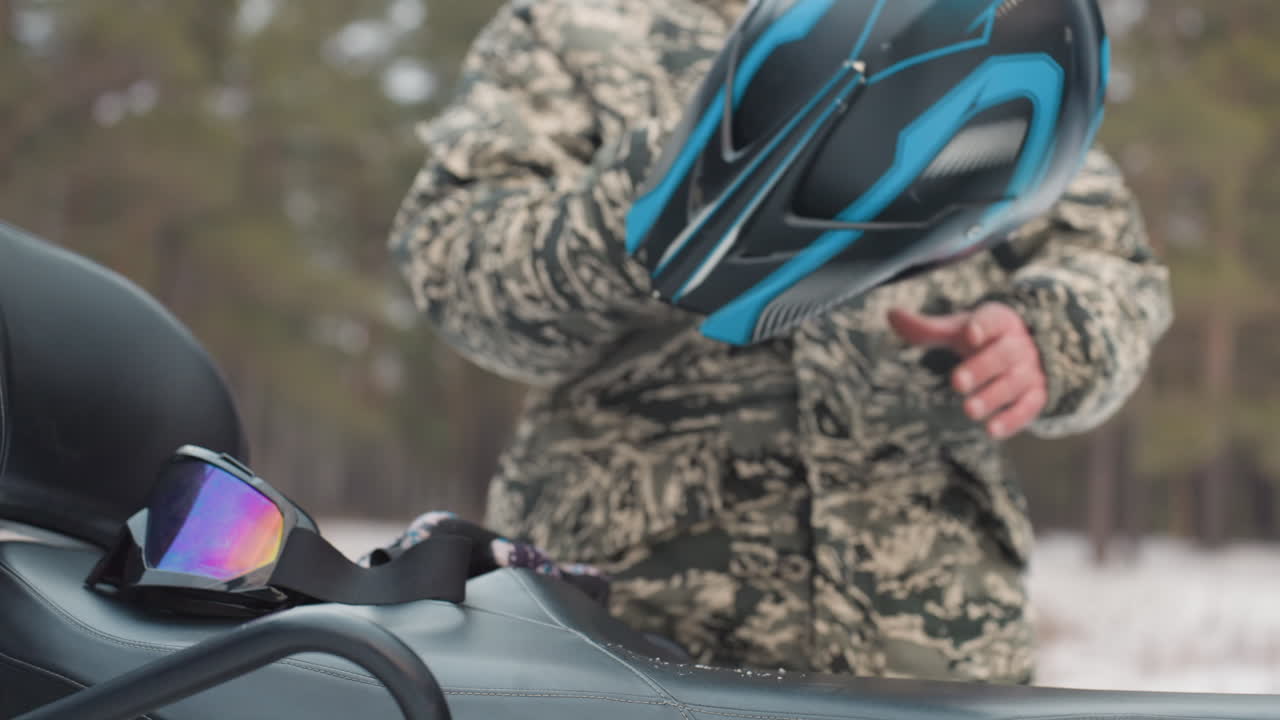 Rider prepares for adventure. Helmet ready before daring journey. Approaching rider gets ready for extreme ride. Camouflage clad rider anticipates intense ATV adventure in winter forest