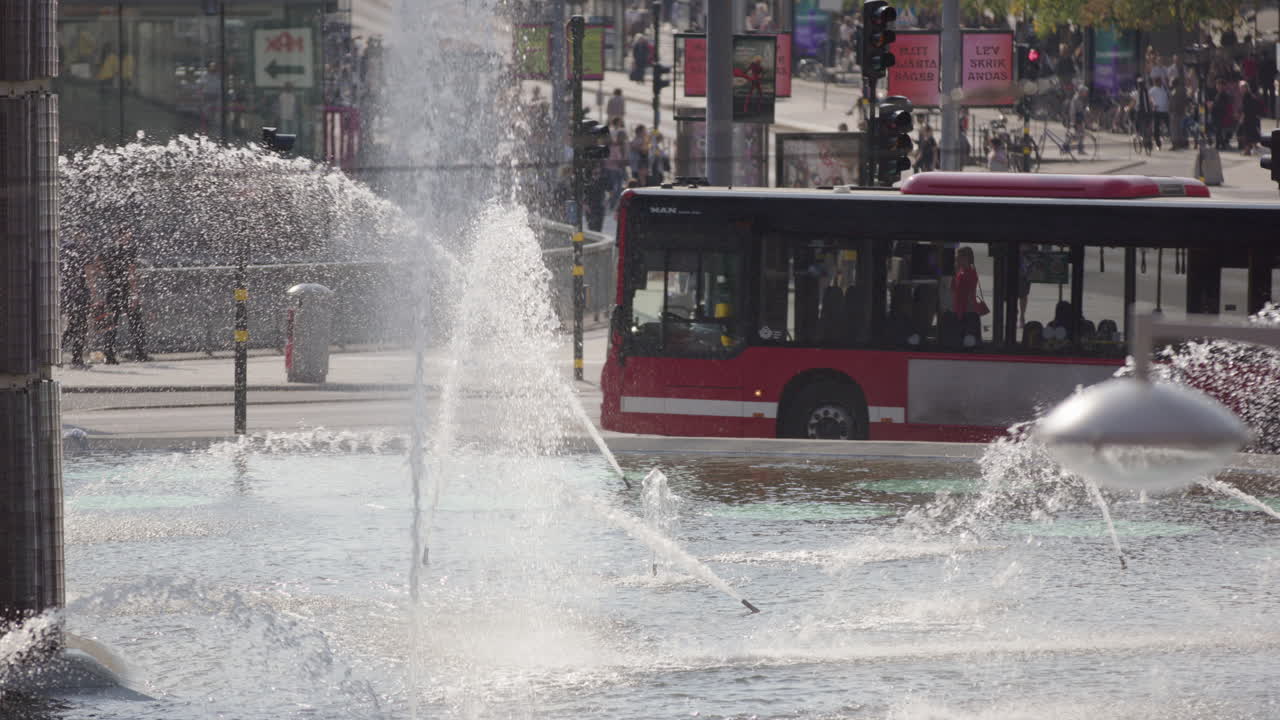 Telephoto slomo view of traffic around fountain spraying water at Sergels Square