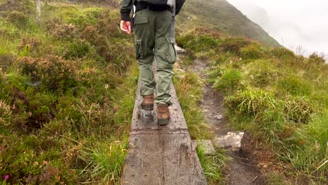 vista trasera en cámara lenta de excursionista con perro caminando por un estrecho sendero de madera en altas montañas verdes de irlanda - día lluvioso con niebla densa en el valle