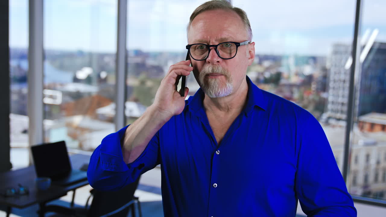 Caucasian adult bearded man in blue shirt and glasses speaks on the phone. Businessman communication with clients and partners in office. Blurred backdrop.