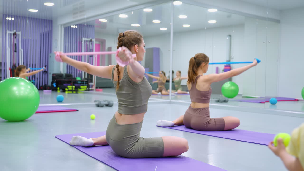 damas participando en una sesión de entrenamiento en grupo, estirando los brazos con bandas de resistencia en esteras púrpuras, con un niño pequeño cerca observando, en un entorno de gimnasio vibrante
