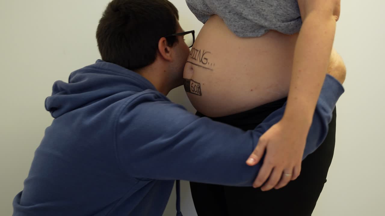 padre abrazando y tocando el vientre de su esposa embarazada con "carga" pintado en él