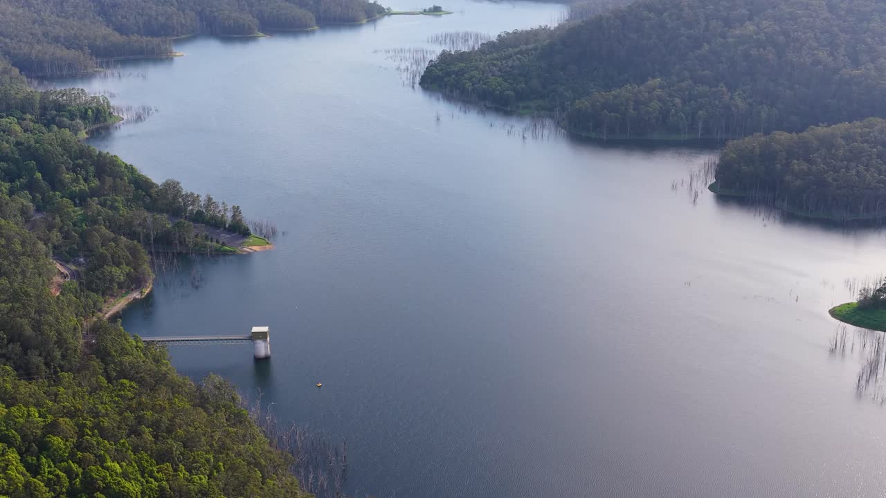 Drone glides over tranquil reservoir, lush forest, and distant hills in soft natural daylight