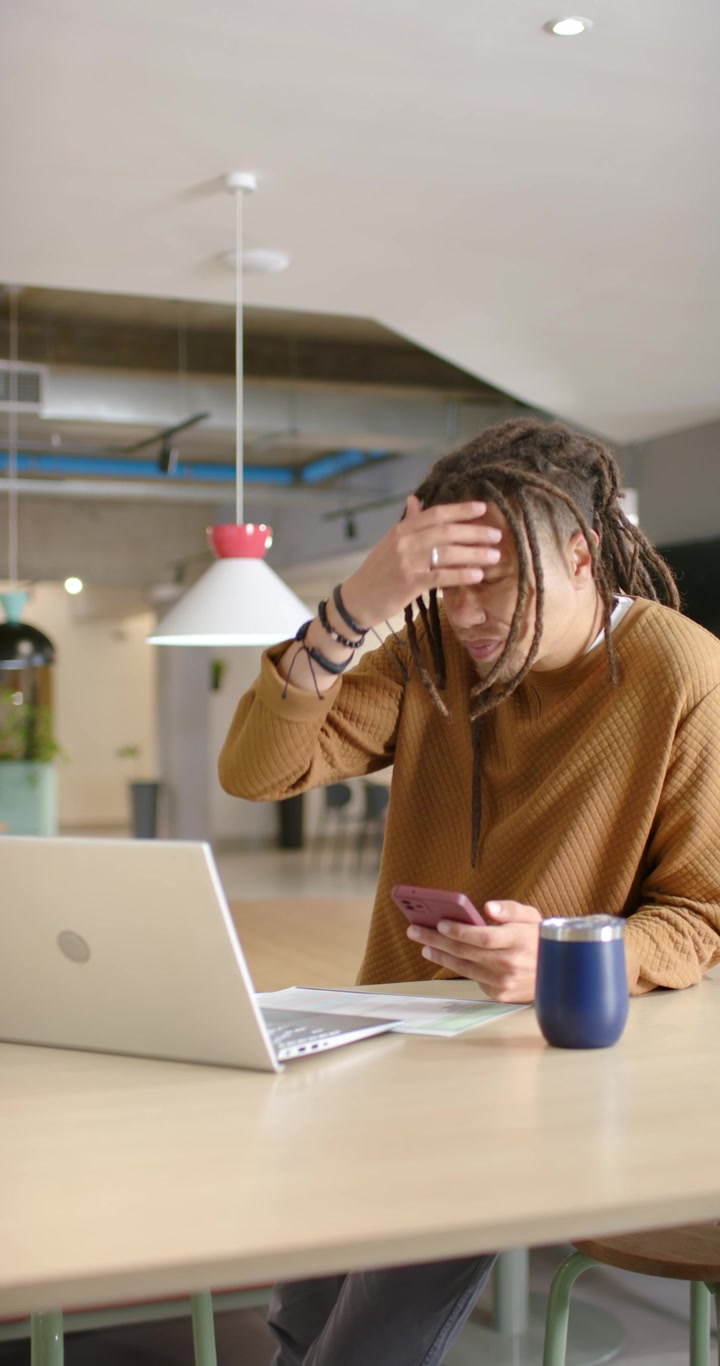 Vertical video: Adult African American man in sweater toggling phone and laptop at desk working
