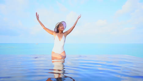 Mientras-Se-Sienta-En-El-Borde-De-Una-Piscina-De-Borde-Infinito,-Una-Atractiva-Joven-En-Traje-De-Baño-Y-Sombrero-Para-El-Sol-Levanta-Los-Brazos-En-Señal-De-Triunfo