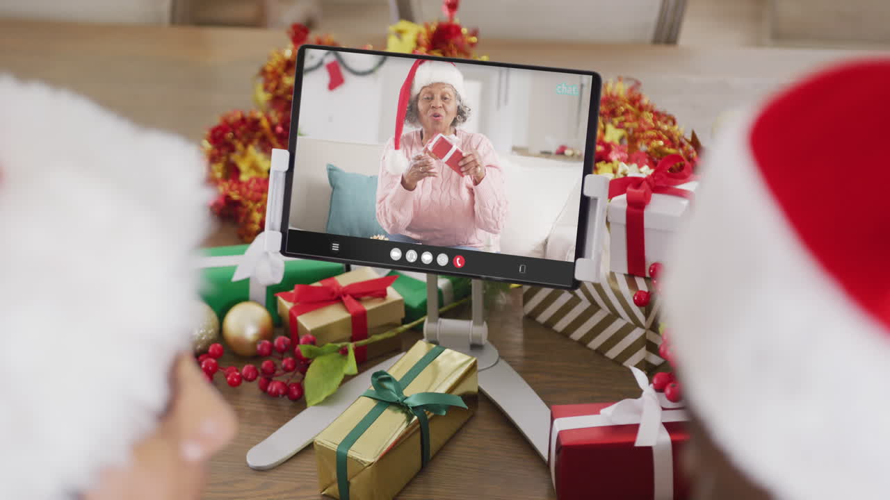 una pareja caucásica con sombreros de santa haciendo una llamada de video de tableta con una feliz mujer afroamericana