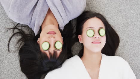 madre biracial feliz y hija adulta en túnicas acostadas con rebanadas de pepino en los ojos, cámara lenta