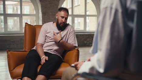 a man with a beard in a pink t-shirt sits on a brown chair and tells a psychologist about his experiences in a brick building with large windows. personal interview during communication with a psychologist