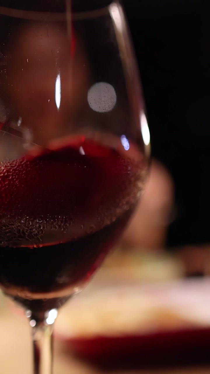 Primo piano di un bicchiere di vino rosso delicatamente roteato su un tavolo da pranzo illuminato caldamente, con una sfocatura morbida dello sfondo e un'atmosfera accogliente e intima.
