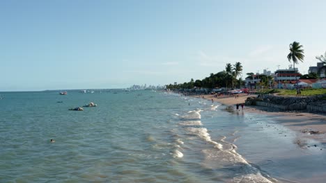 dolly in aerial extreme wide shot of the tropical bessa beach in the capital city of joao pessoa, paraiba, brazil with people enjoying the ocean, small fishings boats and skyscrapers in the background