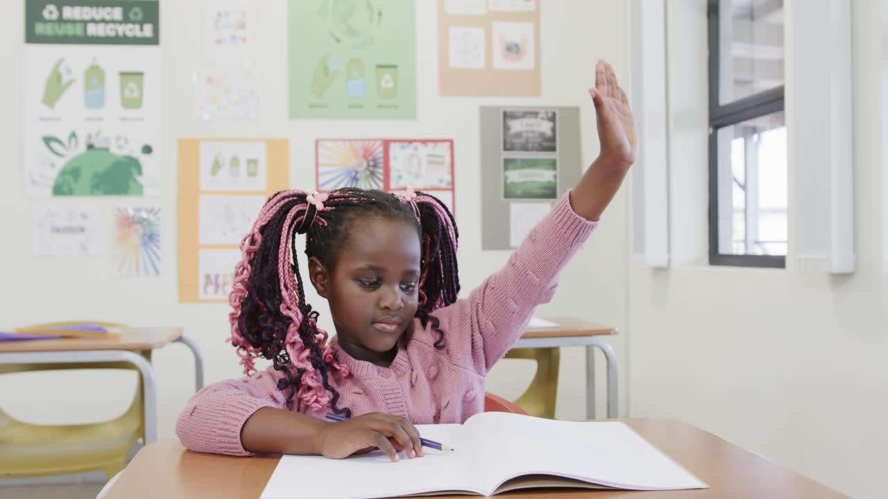 In classroom, African American girl raising hand while writing in ...