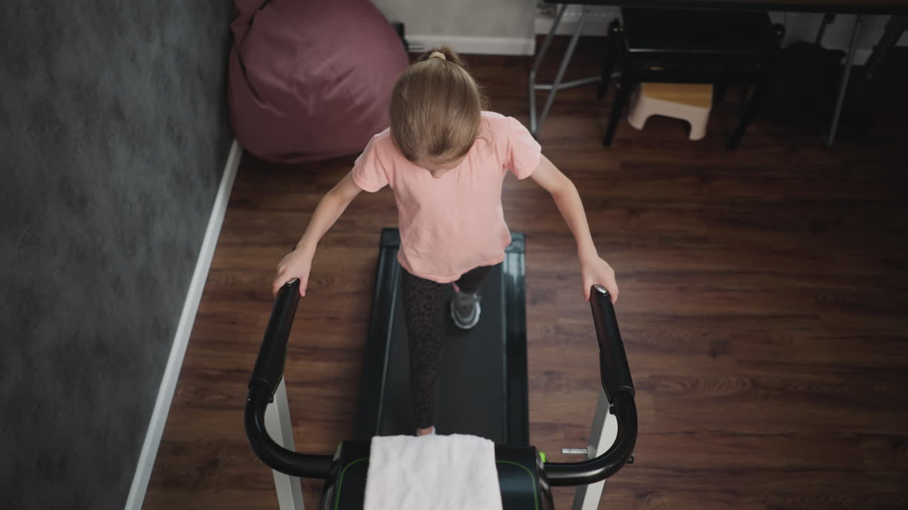 Top down view of fitness lover resting hand on treadmill handle walking steadily on treadmill plugged into power socket with towel draped over console in home gym with grey textured wall wooden floor