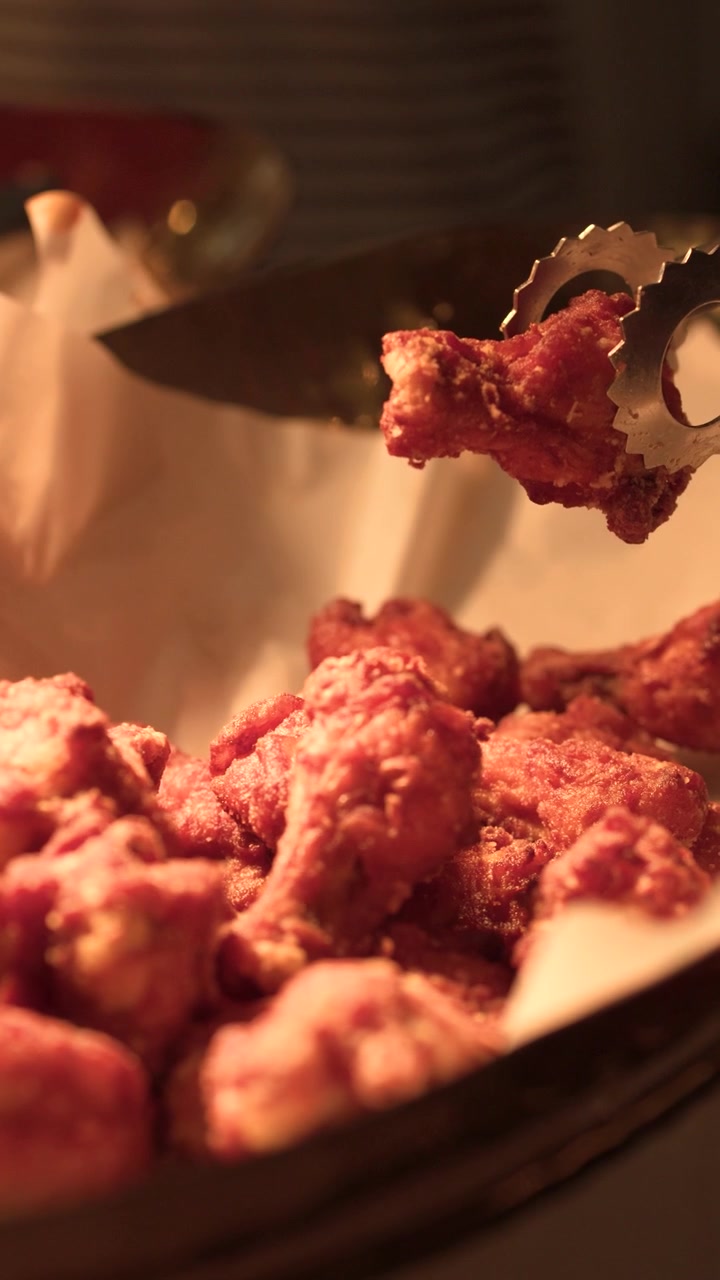 Jepretan close-up penjepit mengangkat ayam goreng pedas dari nampan logam di bawah pencahayaan hangat