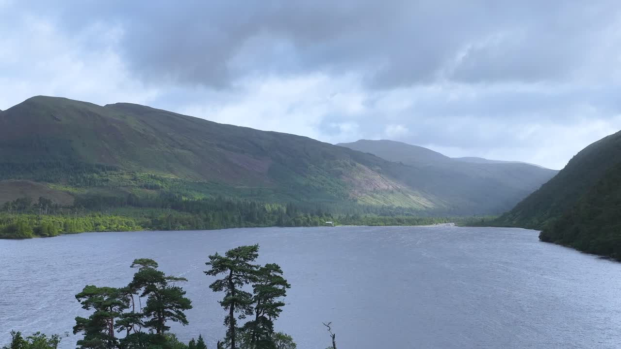 Smooth pan reveals scenic lake, tree-covered island, and misty highland mountains under cloudy daylight