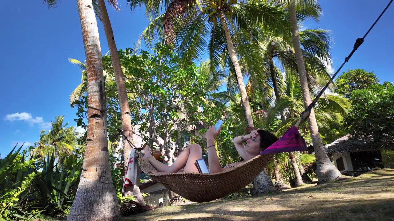 Giovane donna in videochiamata da un'amaca in un resort tropicale sulla spiaggia, circondata da palme.