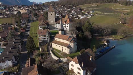 Castillo-De-Spiez-E-Iglesia-Con-El-Lago-Thun-Y-Los-Alpes-Suizos-Al-Fondo,-Día-Despejado,-Vista-Aérea