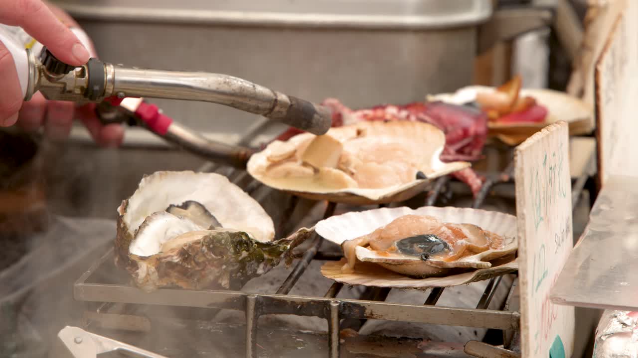 A market vendor uses a blowtorch to sear fresh oysters and scallops on a grill