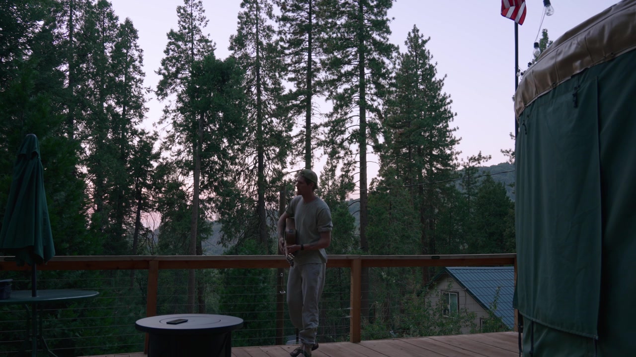 A man walks onto a wooden deck carrying an instrument case, surrounded by pine trees at dusk.