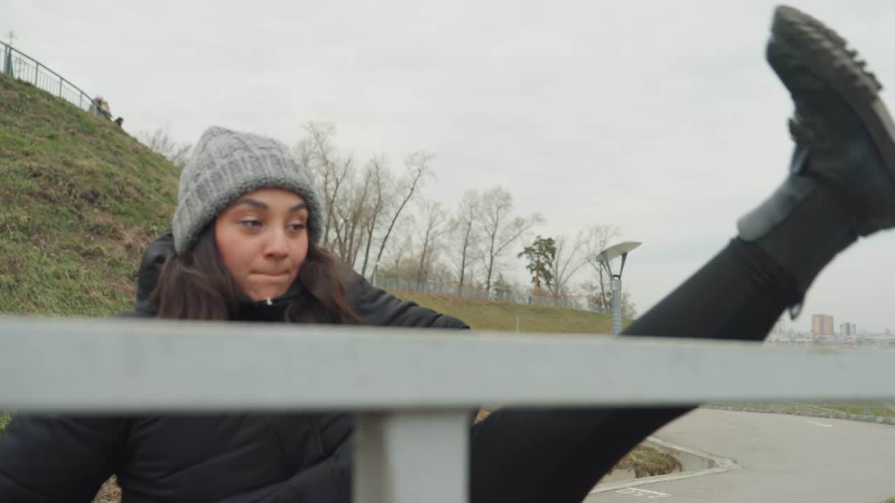 Woman standing by railing warming up, outdoor park path and grassy hill under pale sky, knit hat and puffer jacket, controlled approach into dance warmup with steady reach and step, focus on posture