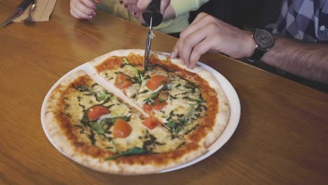 close up of pizza being cut by a man with a pizza cutter into slices seen from above on a wooden table with a woman besides in the background