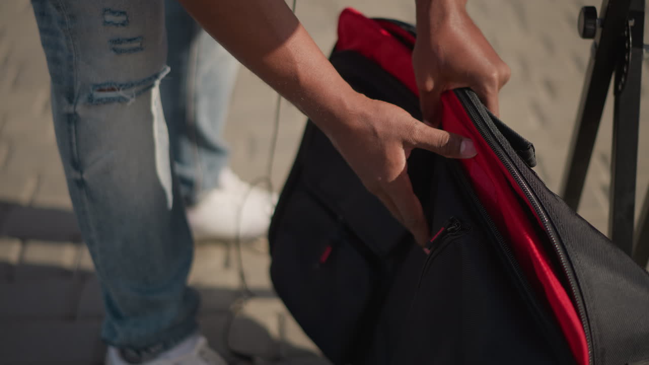 black DJ packing cables into backpack with red interior on sidewalk, unplugging mixer cord and stowing headphones, denim jeans and casual stance, daylight city scene, careful zipper closure, compact