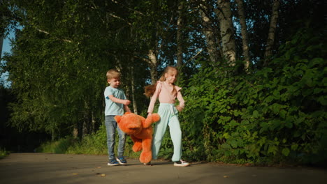 brother and sister walking along forest path on sunny day as girl joyfully marches while holding large orange teddy bear, with boy beside her handing something over, both surrounded by lush greenery
