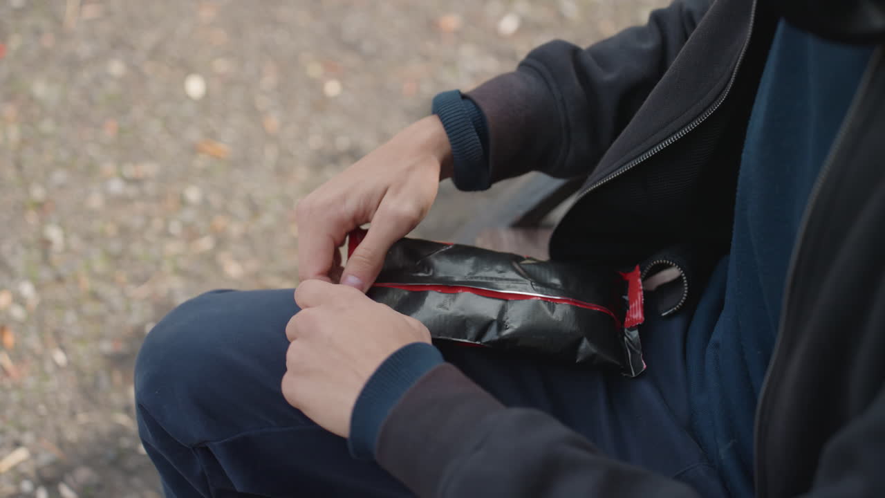 mann på benk åpner godbitpose, fjerner forsiktig liten rød snack fra folieinnpakning, nærbilde som viser glidelås- og jakkedetaljer, urbant fortau og grusbakgrunn, stille fokus på taktil handling