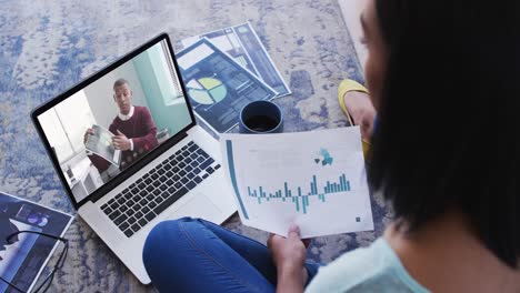 Mixed-race-businesswoman-sitting-on-floor-using-laptop-having-video-call-with-male-colleague