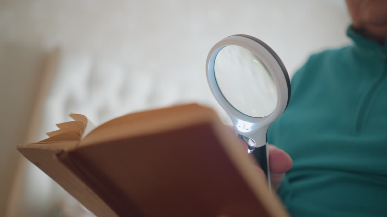 Close-up view of elderly woman reading a book while holding a magnifying glass. Her facial expression suggests focus and slight frustration as she examines the pages carefully