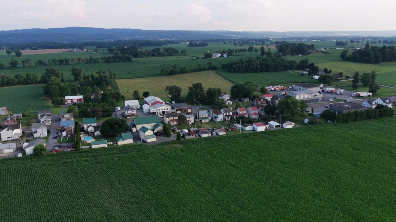 Liten landlig landsby langs en landevei med blandede tak og rød låve, tett grønn maisåker strekker seg mot bølgende jordbruksland og fjerne åser i Pennsylvania, USA. Bredt dronebilde fra luften