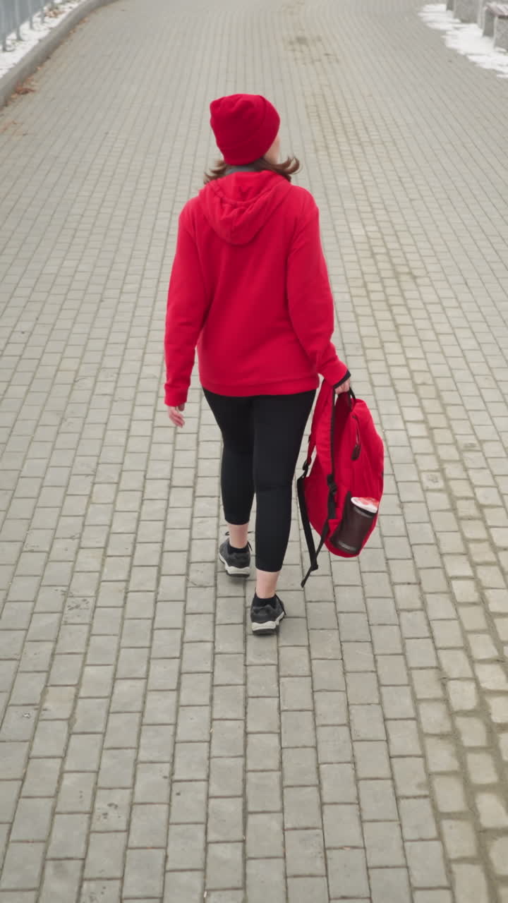 Back view of athlete wearing red winter jacket and head warmer holding backpack in left hand, walking along snowy fog-covered path lined with benches and trees dusted with snow in calm winter setting