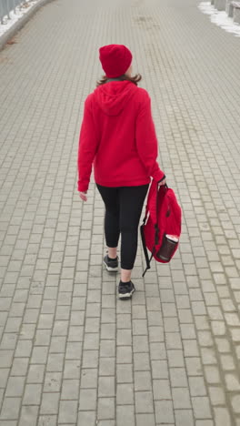back view of athlete wearing red winter jacket and head warmer holding backpack in left hand, walking along snowy fog-covered path lined with benches and trees dusted with snow in calm winter setting