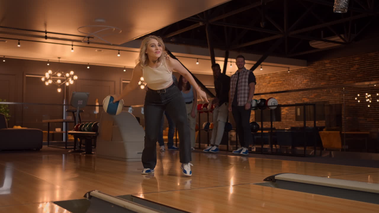 Una giovane donna al bowling lancia una palla sulla pista e cade a terra al rallentatore, poi salta e balla di gioia. Gli amici fan la supportano e applaudono. Amici multietnici giocano a bowling insieme.