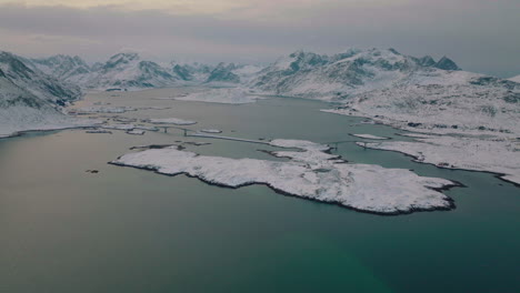 Aerial-view-across-frozen-Ramberg-village-under-glacial-snowy-Lofoten-mountain-range-fjord